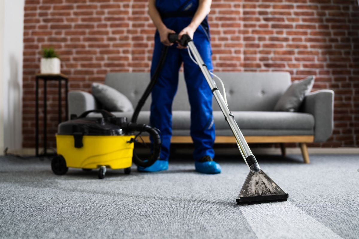A person in blue overalls and shoe covers uses a floor cleaner on a gray carpet, with a yellow vacuum nearby and a brick wall in background.