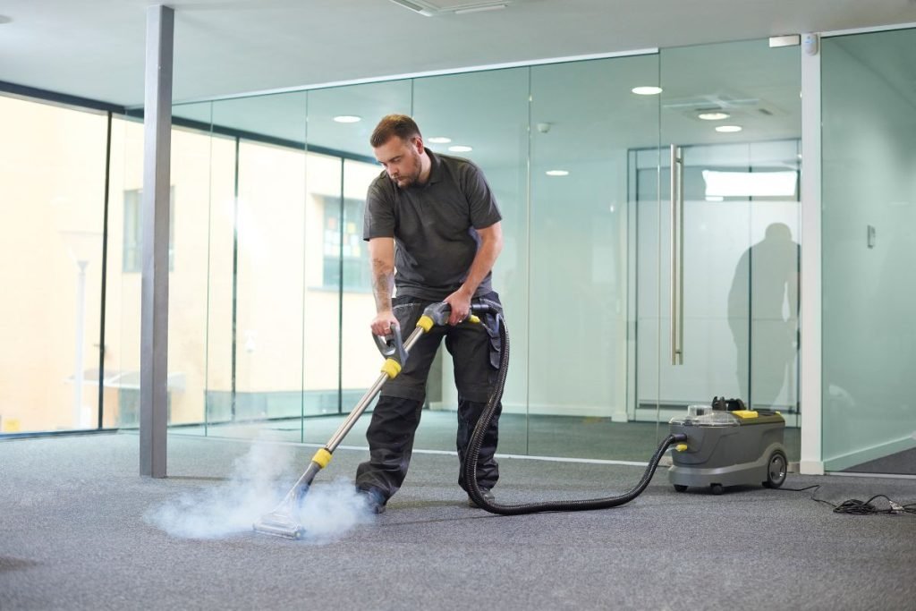 A man using a carpet cleaner in a bright, modern office with large glass windows, producing steam as he works on the floor.