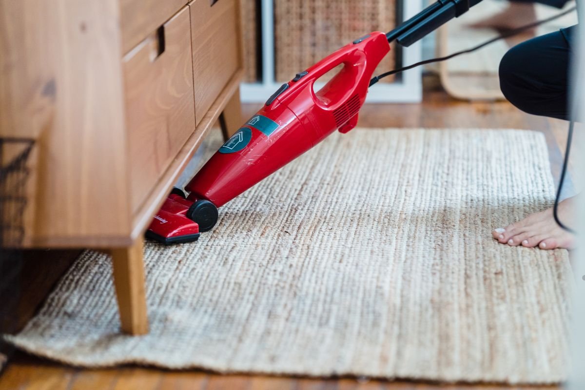 A person using a red vacuum cleaner on a woven rug, reaching under a wooden dresser. A bare foot is visible nearby.