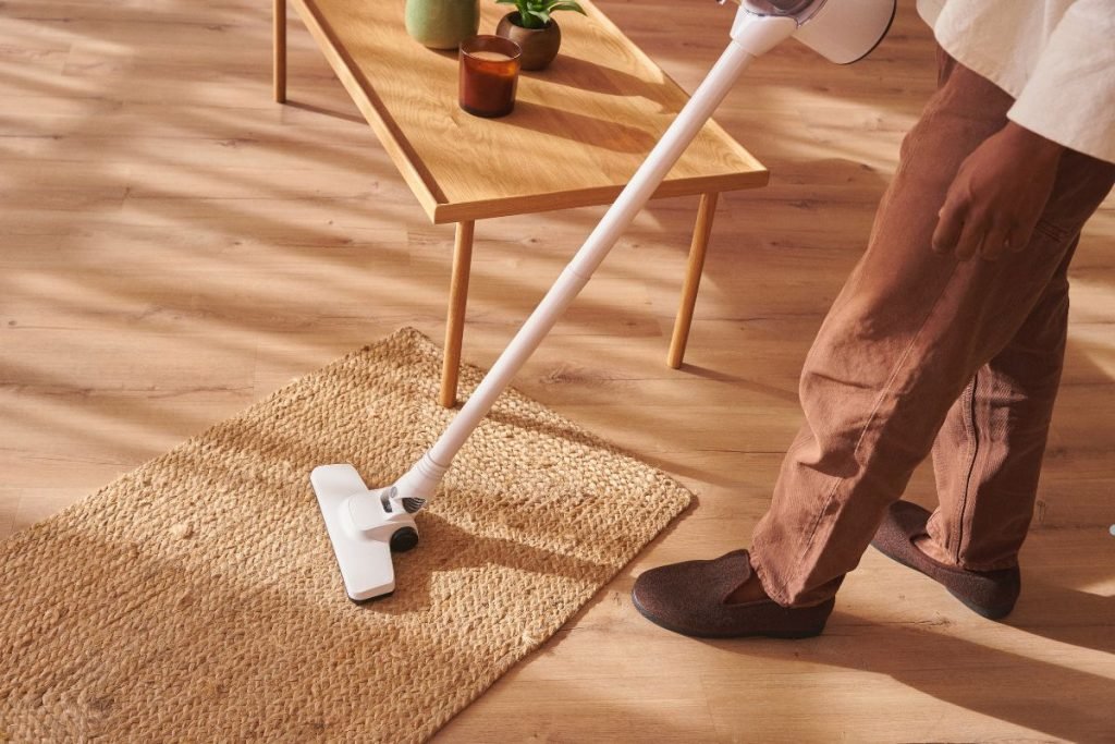 A person vacuuming a woven rug with a cordless vacuum, wearing brown pants and slippers, in a sunlit, cozy living room.