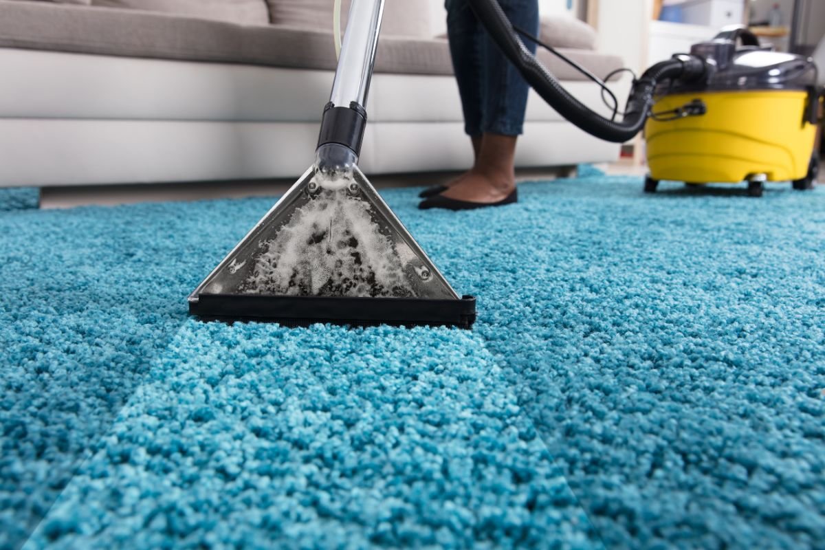 A person cleaning a bright blue carpet with a steam cleaner, revealing suds, while a yellow vacuum cleaner sits nearby.