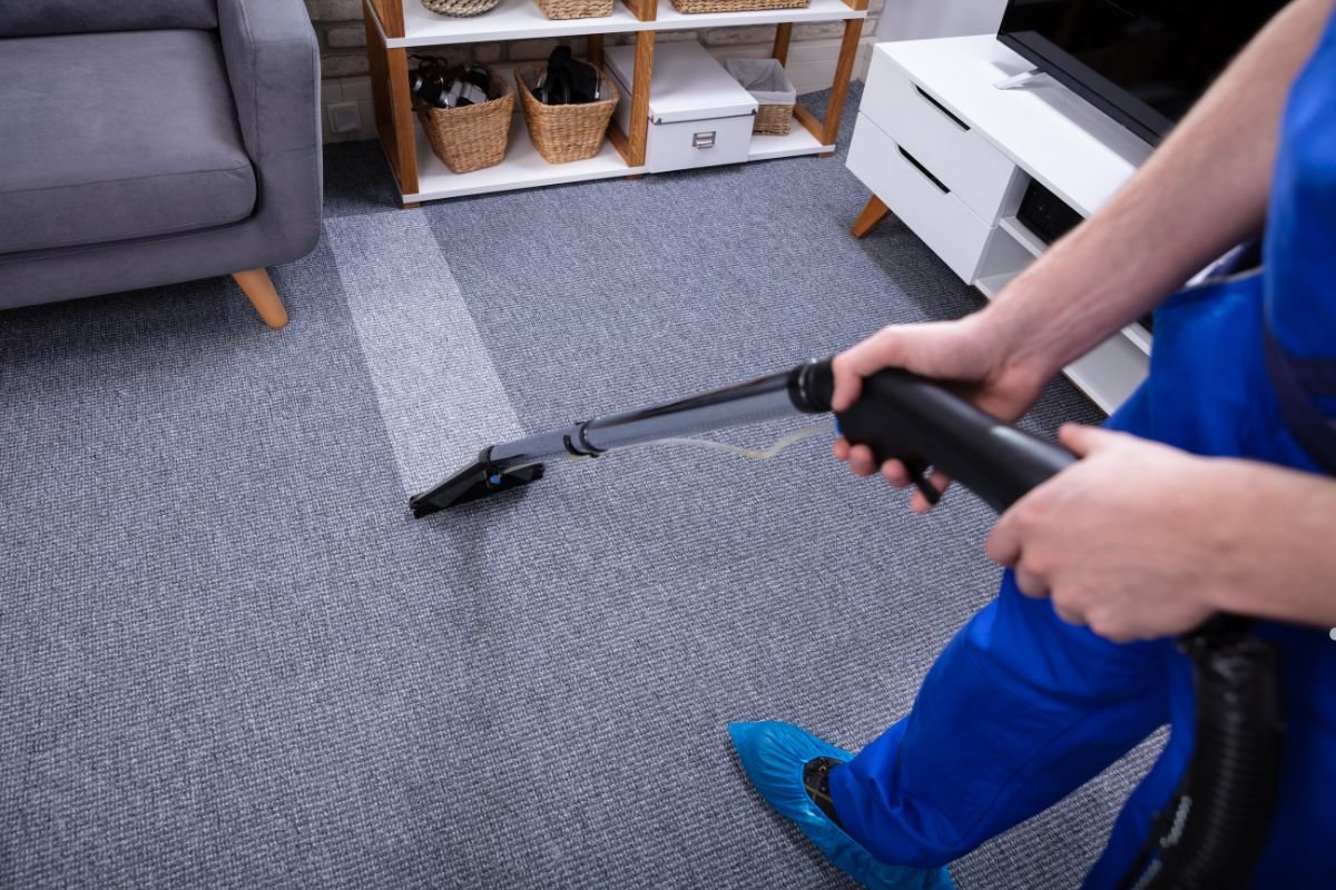 A person in blue cleaning attire uses a carpet cleaner on a gray carpet, with a clean strip visible among the uncleaned area.