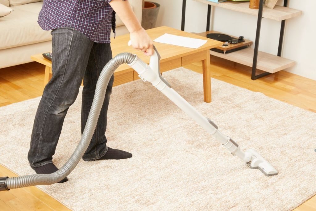 A person in jeans and a checkered shirt vacuums a carpeted living room with a modern vacuum cleaner, showcasing a tidy home environment.